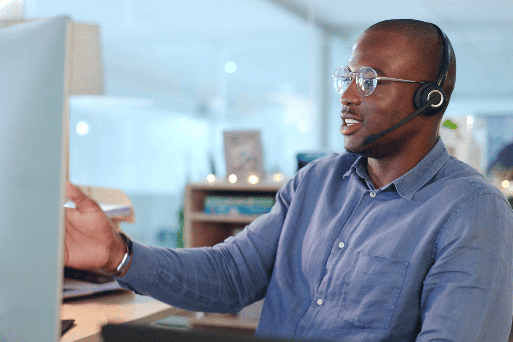 A man sat infront of a oomputer monitor talking on a headset